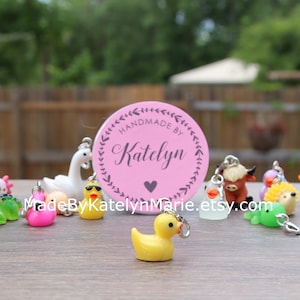 May include: A collection of colorful animal charms, including a pink flamingo, a green frog, a yellow duck, a white swan, a brown cow, a white chicken, and a brown hedgehog. The charms are handmade and feature a pink circle with the text "Handmade by Katelyn" and a pink heart.