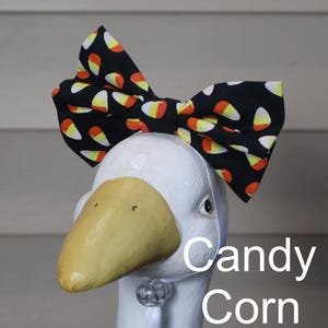 May include: A white goose head sculpture with a large black bow tie featuring a candy corn pattern. The bow tie has a repeating design of orange, yellow, and white candy corn shapes on a black background. The text "Candy Corn" is below the goose's head.