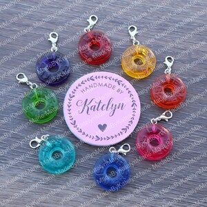 May include: Assortment of colorful, handmade, candy-shaped, plastic stitch markers with silver lobster clasps. The stitch markers are arranged around a pink circle with the text "HANDMADE BY Katelyn" in black lettering.