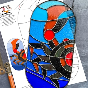 May include: A stained glass window hanging from a chain. The window is oval shaped and features a colorful abstract design with blue, red, black, and orange glass pieces. The window is surrounded by a black metal frame.