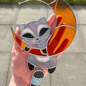 May include: A stained glass window hanging features a raccoon with a long, striped tail hanging from a crescent moon. The moon is orange and red with a silver outline. The raccoon is gray and white with black eyes and a black nose.