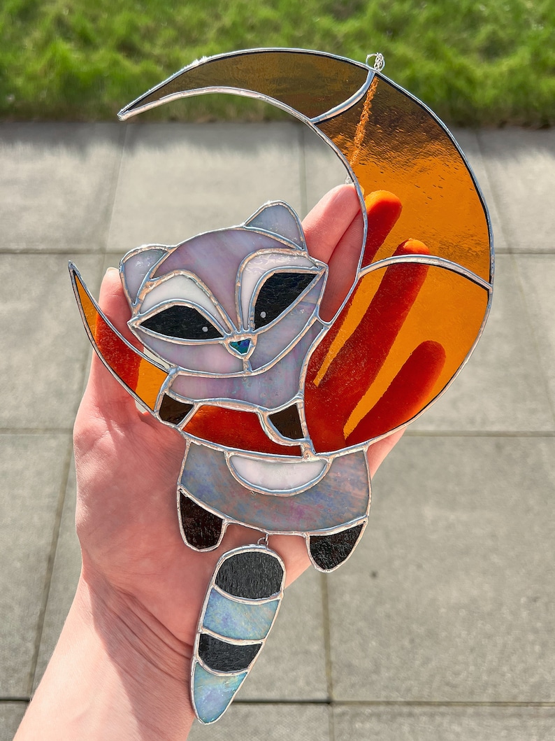 May include: A stained glass window hanging features a raccoon with a long, striped tail hanging from a crescent moon. The moon is orange and red with a silver outline. The raccoon is gray and white with black eyes and a black nose.