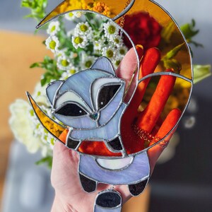 May include: A stained glass window hanging features a purple raccoon with a black tail hanging from a crescent moon with a yellow and orange gradient.
