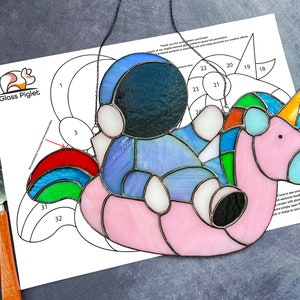 Könnte beinhalten: Ein Bleiglasmuster für ein Einhorn, das einen Astronauten auf einem rosa aufblasbaren Ring reitet. Das Muster ist nummeriert und enthält eine schwarz-weiße Umrandung des Designs.