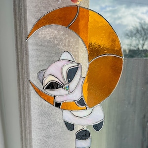 May include: A stained glass suncatcher featuring a cartoon raccoon hanging from a crescent moon. The moon is made of orange glass and the raccoon is white with black accents.