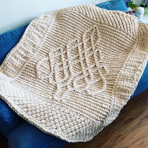 Könnte beinhalten: Eine cremefarbene gestrickte Wolldecke mit einem großen, komplizierten gewebten Muster in der Mitte. Die Decke liegt über einem blauen Sofa.