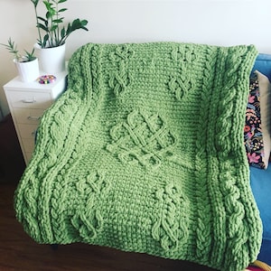 May include: A green knitted throw blanket with a cable knit pattern. The blanket has a large, intricate design in the center and is draped over a couch.