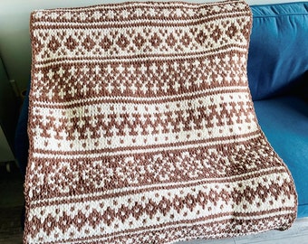 May include: A brown and white knitted throw blanket with a geometric pattern. The blanket is folded and placed on a sofa.