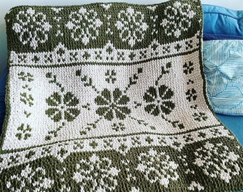 May include: A green and white knitted throw blanket with a floral pattern. The blanket is folded and placed on a blue sofa.