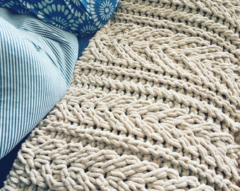 Pode incluir: Um cobertor bege de malha grossa com um padrão tecido texturizado. Duas almofadas são visíveis: uma com um desenho floral azul e branco, e outra com riscas azuis e brancas. O cobertor parece macio e aconchegante.