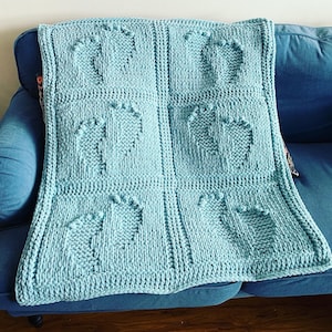 Peut inclure: Une couverture pour bébé en tricot bleu clair avec un motif répétitif de petits cercles. La couverture est pliée et placée sur un canapé bleu.