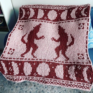 May include: A knitted blanket in shades of pink and brown. The blanket features a repeating pattern of stylized figures and moon phases. The figures are in a dancing pose. The blanket is folded over a blue surface.