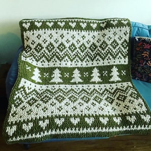 May include: A green and white knitted throw blanket with a traditional Scandinavian pattern featuring stylized trees. The blanket is folded and placed on a couch.