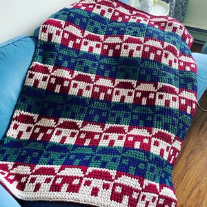 Könnte beinhalten: Eine gehäkelte Wolldecke mit einem sich wiederholenden Muster aus roten, weißen, grünen und blauen Häusern. Die Decke ist aus dickem Garn gefertigt und hat eine weiche, strukturierte Haptik.