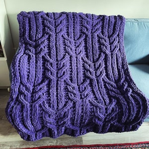 May include: A purple knitted throw blanket with a cable knit pattern. The blanket is soft and fluffy and would be perfect for cuddling up on the couch.