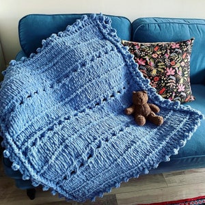 May include: A blue knitted throw blanket with a textured pattern and a pom-pom border. A brown teddy bear is sitting on the blanket.