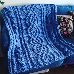 May include: A blue knitted throw blanket with a cable knit pattern. The blanket is draped over a blue sofa.