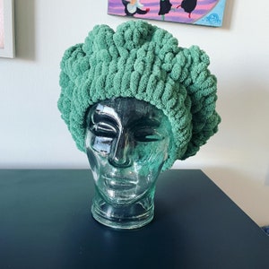May include: A green crocheted hat on a glass mannequin head. The hat is made of thick yarn and has a textured, fluffy appearance.