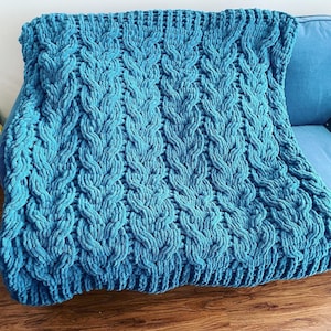 May include: A teal blue, chunky knit blanket with a cable knit design. The blanket is made of a soft, plush material and is draped over a blue sofa. The texture of the blanket is visible, with the raised pattern creating a cozy look.