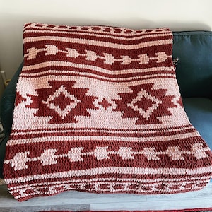 Könnte beinhalten: Eine rot- und rosafarbene gestrickte Wolldecke mit geometrischem Muster. Die Decke ist gefaltet und über ein Sofa gelegt.