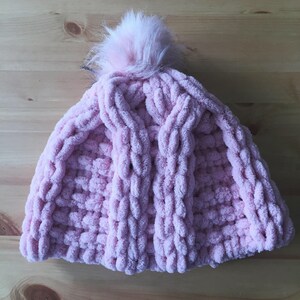 May include: A pink knitted hat with a fluffy white pom pom on top. The hat is made with a chunky yarn and has a cable knit pattern.