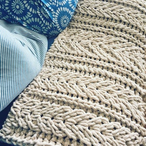 May include: A beige, chunky knit blanket with a textured, woven pattern. Two pillows are visible: one with a blue and white floral design, and another with blue and white stripes. The blanket appears soft and cosy.