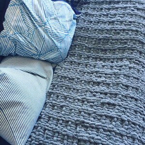 May include: A soft, blue, chunky knit throw blanket with a textured weave. The blanket is folded and placed on a bed with two blue and white patterned pillows.