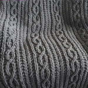 May include: A gray chunky knit blanket with a cable knit pattern. The blanket is soft and fluffy and would be perfect for cuddling up on the couch.