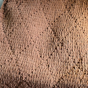 May include: A large, brown, chunky knit blanket with a diamond pattern. The blanket is draped over a couch, showcasing its texture and warmth. The color is a muted, earthy tone, suitable for home decor.