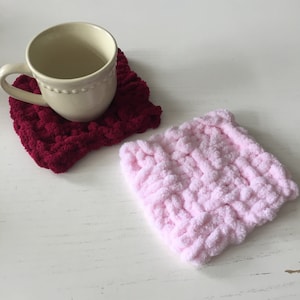 Puede incluir: Dos posavasos de punto, uno rosa y otro burdeos, con una taza de cerámica blanca sobre el posavasos burdeos.