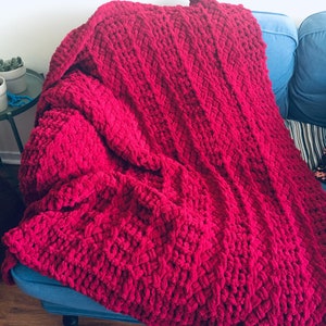 Puede incluir: Una manta de punto de color rojo oscuro con un patrón de punto de cable grueso. La manta está drapeada sobre un sofá azul.