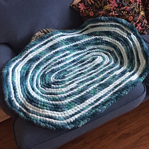 May include: A large oval rug made of chunky yarn in shades of blue, green, and white. The rug is hand-knitted and has a textured, fluffy surface.