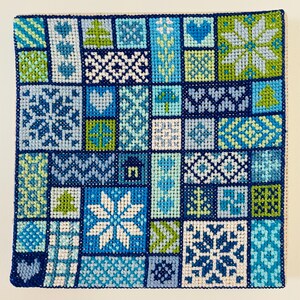 Op de afbeelding: Een blauwe en groene kruissteek sampler met verschillende winterse ontwerpen, waaronder sneeuwvlokken, bomen en harten.