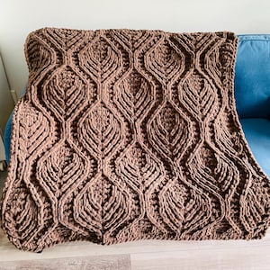 May include: A brown knitted throw blanket with a geometric pattern. The blanket is made of a soft, fluffy yarn and has a textured surface.