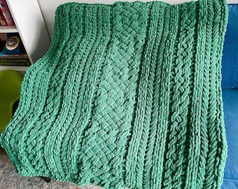 May include: A green knitted throw blanket with a cable knit pattern. The blanket is made of soft, chunky yarn and has a textured surface.