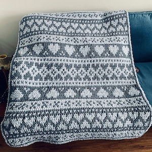 May include: A gray and white knitted throw blanket with a geometric pattern of hearts and snowflakes. The blanket is folded and draped over a couch.