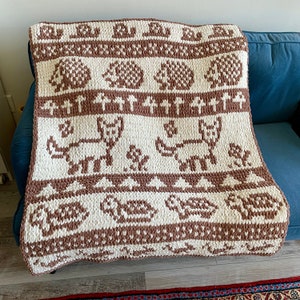 Peut inclure: Une couverture en tricot marron et blanc avec un motif répété d'animaux, y compris des chats, des cerfs et des tortues. La couverture est faite avec un fil épais et a une texture douce et confortable.