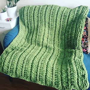 Puede incluir: Una manta de punto verde con un patrón de punto de cable grueso. La manta está drapeada sobre un sillón azul.