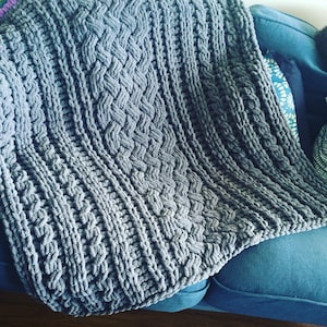 May include: A gray knitted throw blanket with a braided pattern. The blanket is draped over a blue couch.