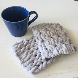 May include: Two gray knitted coasters made of chunky yarn. The coasters are soft and fluffy and would be perfect for protecting surfaces from hot mugs or cups.