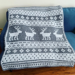 May include: A gray and white knitted throw blanket with a reindeer pattern. The blanket is folded and draped over a blue couch.
