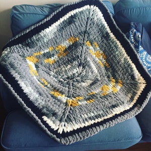 Puede incluir: Una manta de crochet gris, blanco y amarillo con forma cuadrada y diseño texturizado.