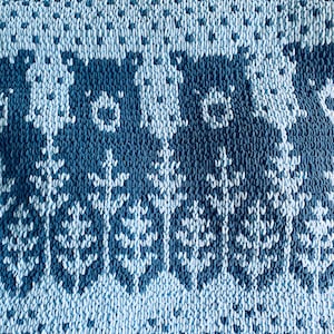 Könnte beinhalten: Nahaufnahme eines Stricktextils mit einem sich wiederholenden Muster aus dunkelblauen Bären und Bäumen auf hellblauem Hintergrund. Der Stoff hat ein strukturiertes Aussehen, mit sichtbaren Stichen, die das Design bilden.