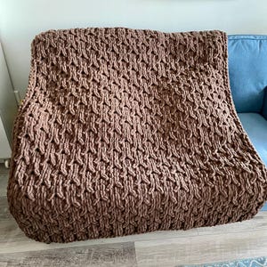 May include: A brown knitted throw blanket with a textured pattern. The blanket is folded and placed on a couch.