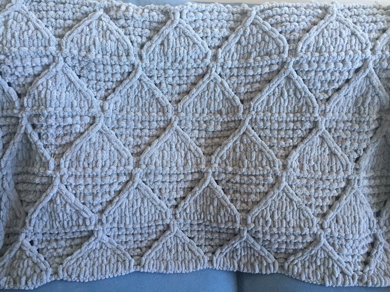 May include: A soft, gray knitted throw blanket with a diamond pattern. The blanket is made with a fluffy yarn and has a textured surface.