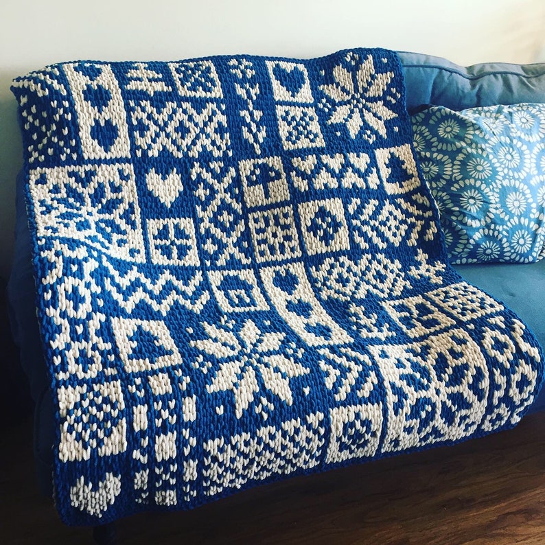 May include: A blue and white knitted blanket with a geometric pattern of squares, hearts, and snowflake-like designs. The blanket is draped over a blue couch, showcasing its intricate details and cozy texture. A matching pillow is visible.