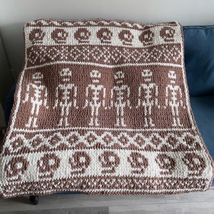 May include: A brown and white knitted throw blanket with a pattern of skeletons and skulls. The blanket is made of chunky yarn and has a soft, cozy texture.