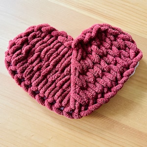 May include: A heart-shaped knitted throw pillow in a deep burgundy color. The pillow is made with a chunky yarn and has a textured, woven design.