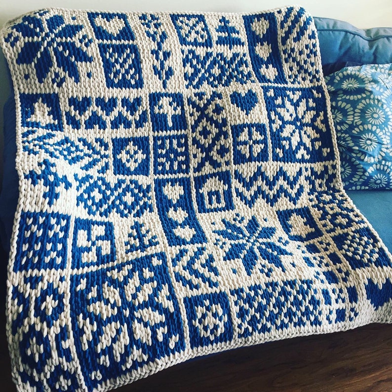 May include: A blue and white knitted blanket with a geometric pattern. The blanket features a grid of squares, each containing a different design, including hearts, snowflakes, and geometric shapes. The blanket is folded over a blue surface.