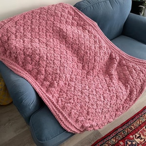 May include: A dusty rose-colored, handmade crochet blanket with a textured, raised pattern. The blanket is draped over a blue armchair. The crochet stitch creates a dense, plush surface, perfect for adding warmth and comfort to any space.